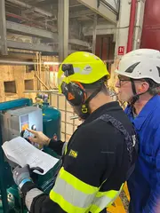 Two technicians wearing safety helmets and hearing protection operating and inspecting an industrial oil filtration unit.